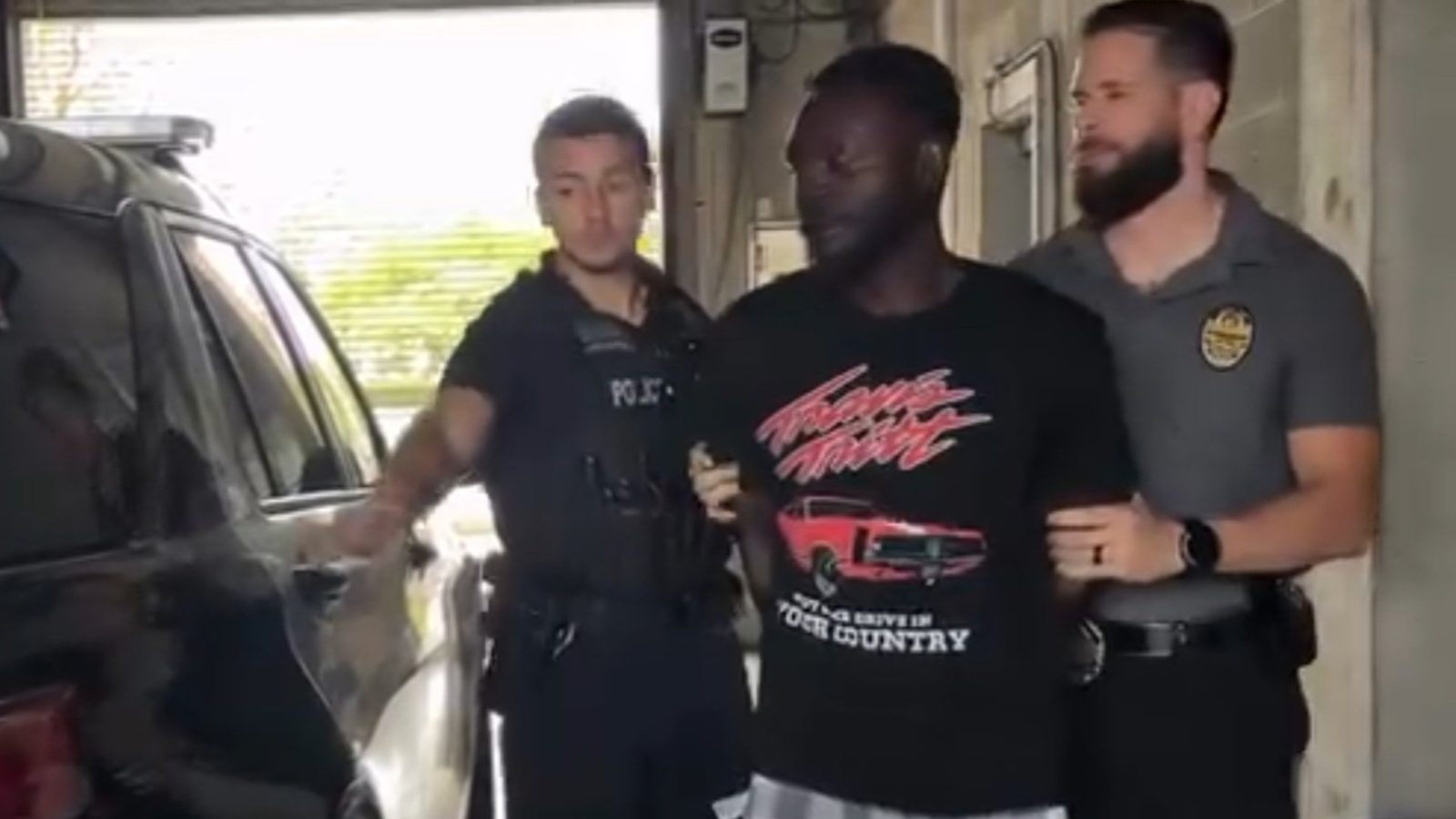 Police officers escorting a man in handcuffs towards a police vehicle in a garage setting.