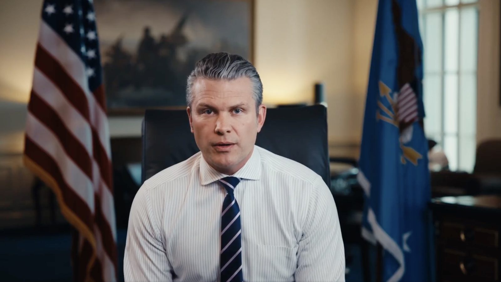 Man in a striped shirt and tie sits in an office with American flags in the background, delivering an address.