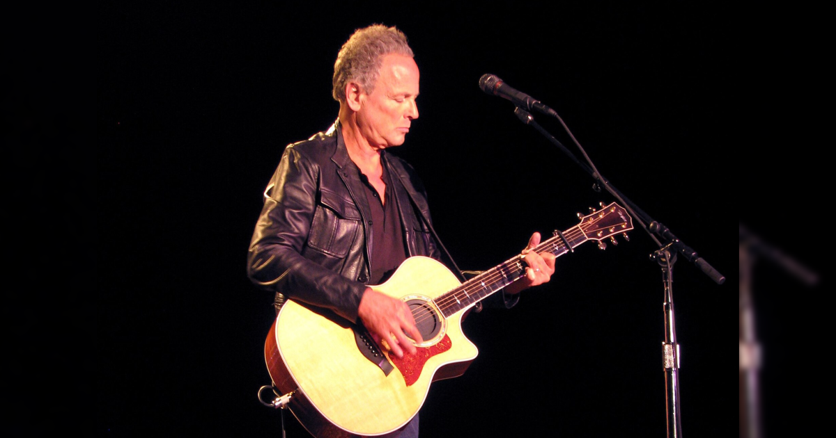 Musician performing on stage with an acoustic guitar, wearing a black leather jacket, captured in a spotlight against a dark background.