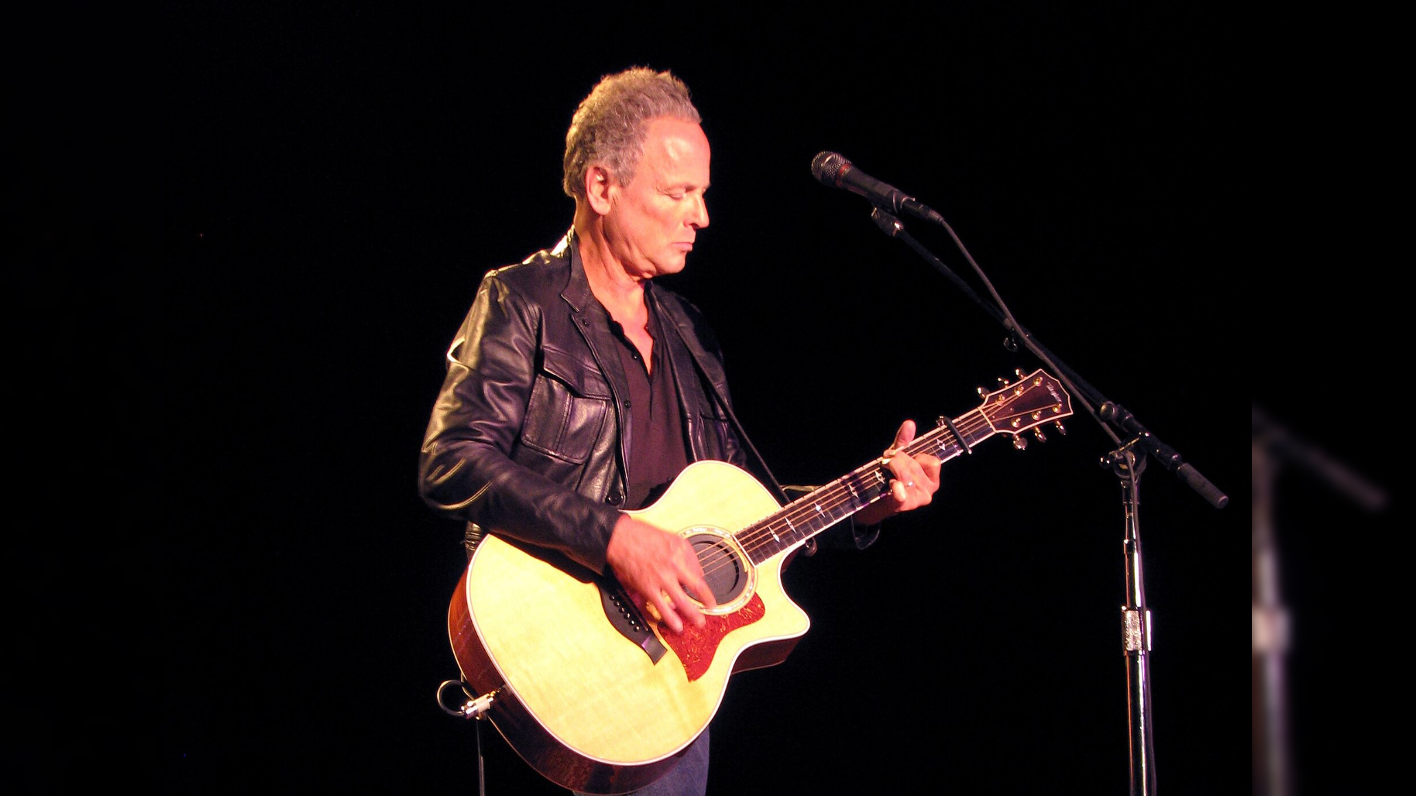 Musician performing on stage with an acoustic guitar, wearing a black leather jacket, captured in a spotlight against a dark background.