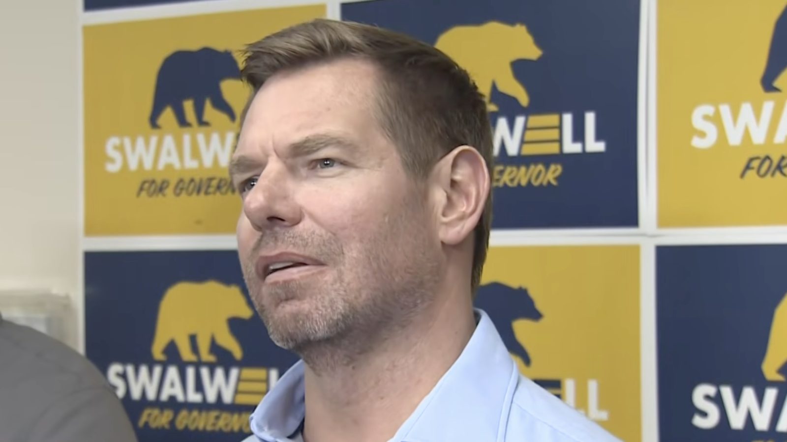 Man speaking at a campaign event with "Swalwell for Governor" banners in the background, emphasizing political engagement and candidacy.