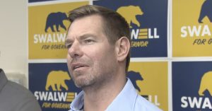 Man speaking at a campaign event with "Swalwell for Governor" banners in the background, emphasizing political engagement and candidacy.