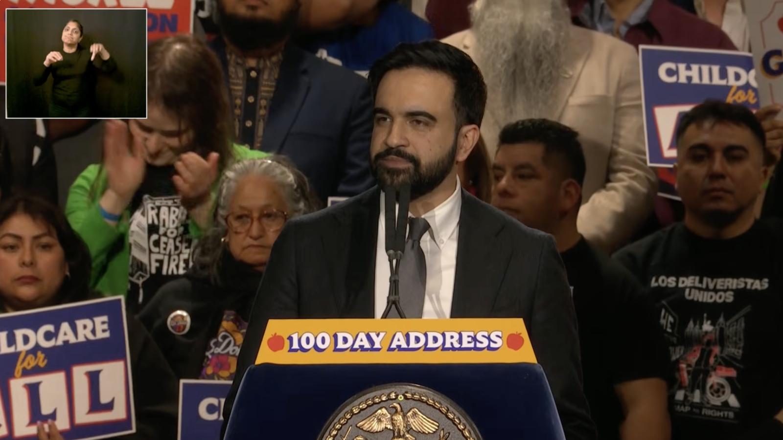 Speaker addressing the audience at a community event, with supporters holding signs advocating for childcare and social issues in the background.