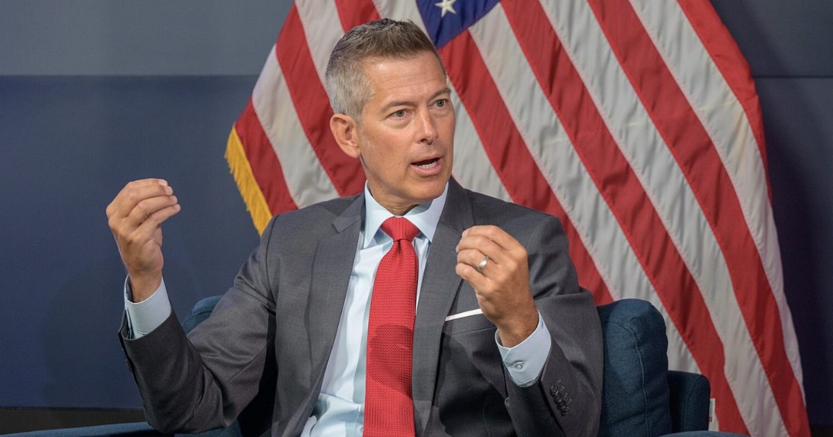 Man in a suit gestures while speaking, with an American flag in the background, during a discussion or interview setting.