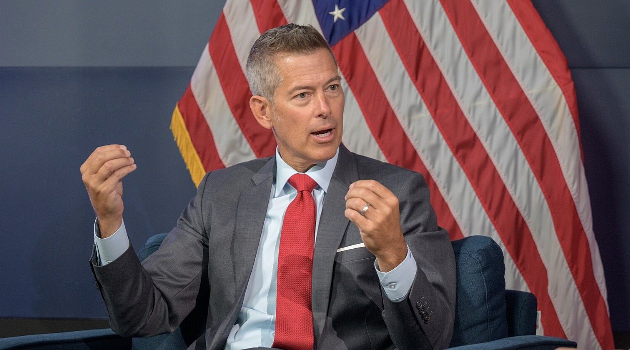 Man in a suit gestures while speaking, with an American flag in the background, during a discussion or interview setting.