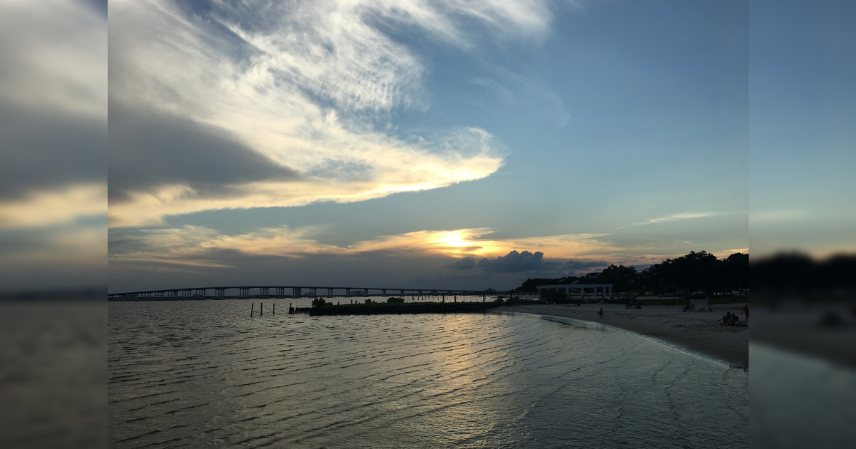 Scenic sunset over a calm beach with gentle waves, a bridge in the background, and people enjoying the shoreline.