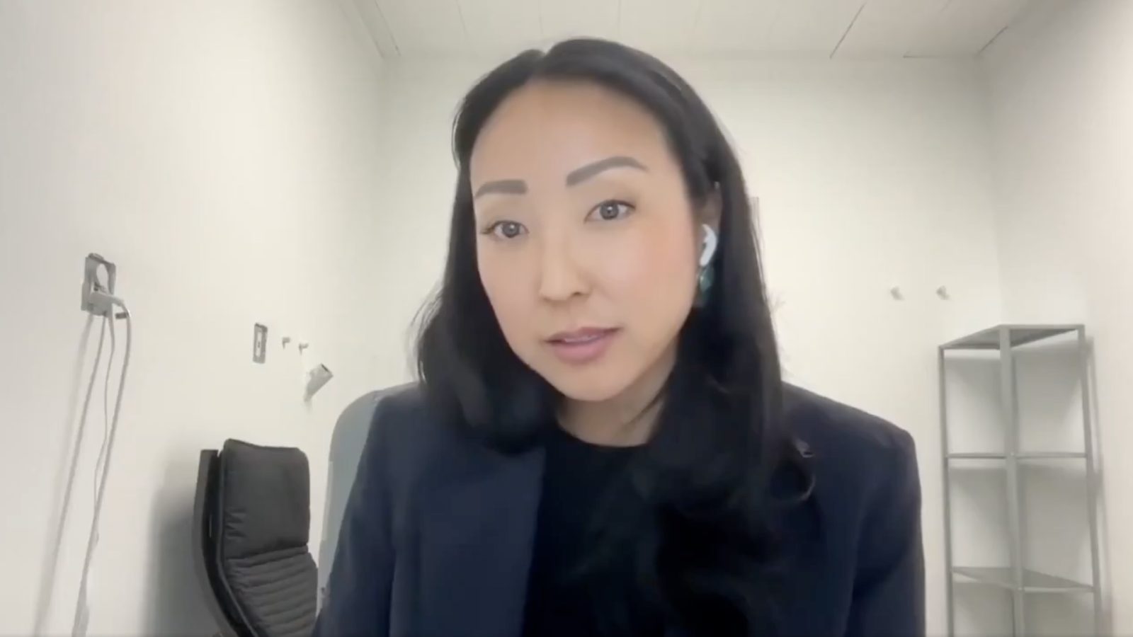 Professional woman speaking in a modern office setting, wearing earbuds and a dark blazer, with a minimalistic background.