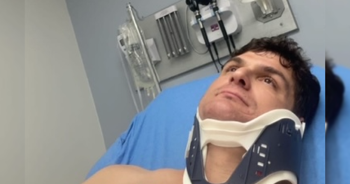 Man in a neck brace lying in a hospital bed, looking up with medical equipment in the background.