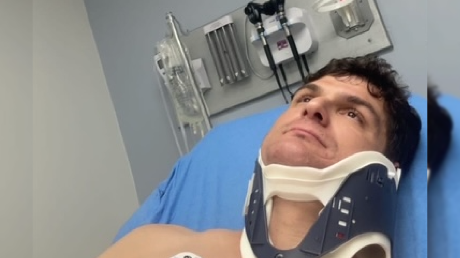 Man in a neck brace lying in a hospital bed, looking up with medical equipment in the background.