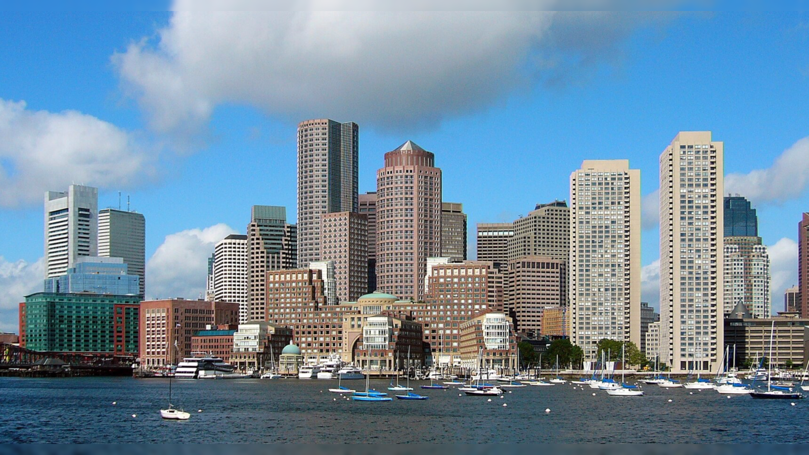 Boston skyline featuring modern skyscrapers and boats in the harbor under a clear blue sky.