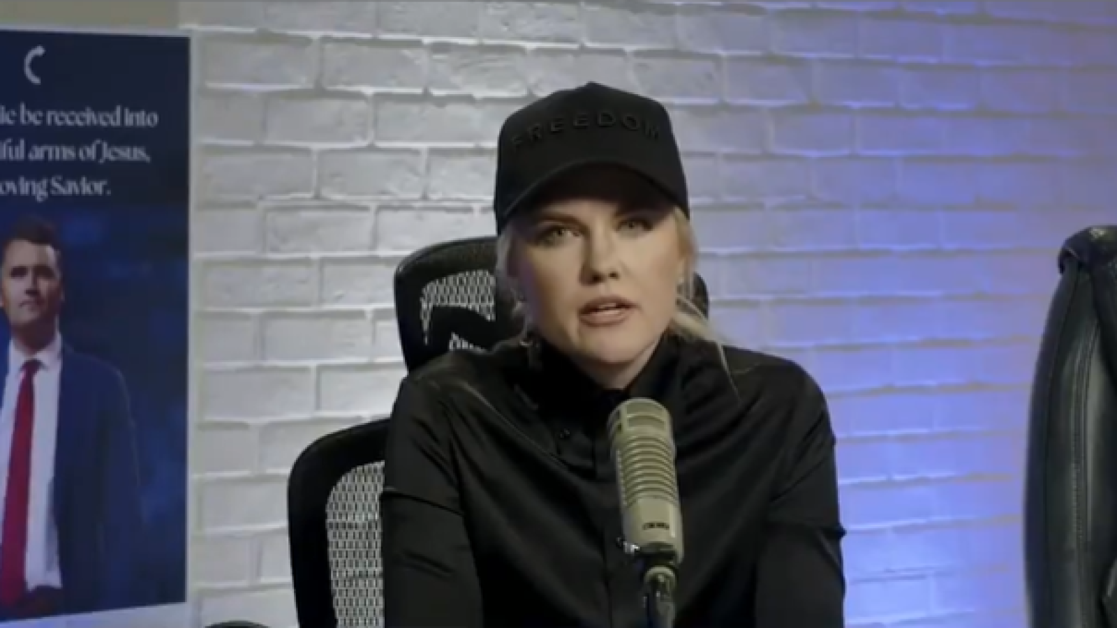 Woman wearing a black cap with the word "FREEDOM" speaks into a microphone during a podcast, with a poster of a man in a suit in the background.