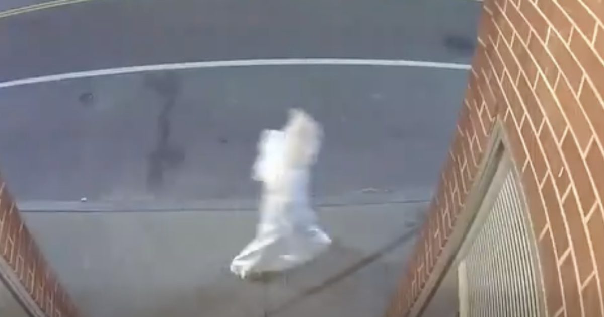 A person in a white outfit walks along the sidewalk near a brick building, captured by a security camera.