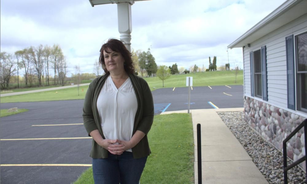 Woman standing outdoors near a building, with green grass and a parking lot in the background, on a cloudy day.