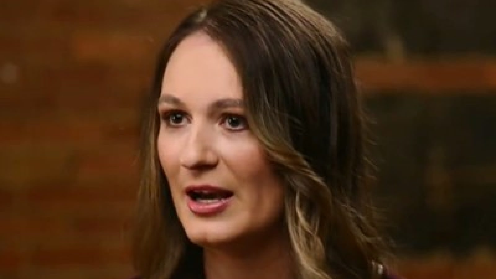 Portrait of a woman with long brown hair speaking in front of a rustic brick background, conveying emotion and engagement.
