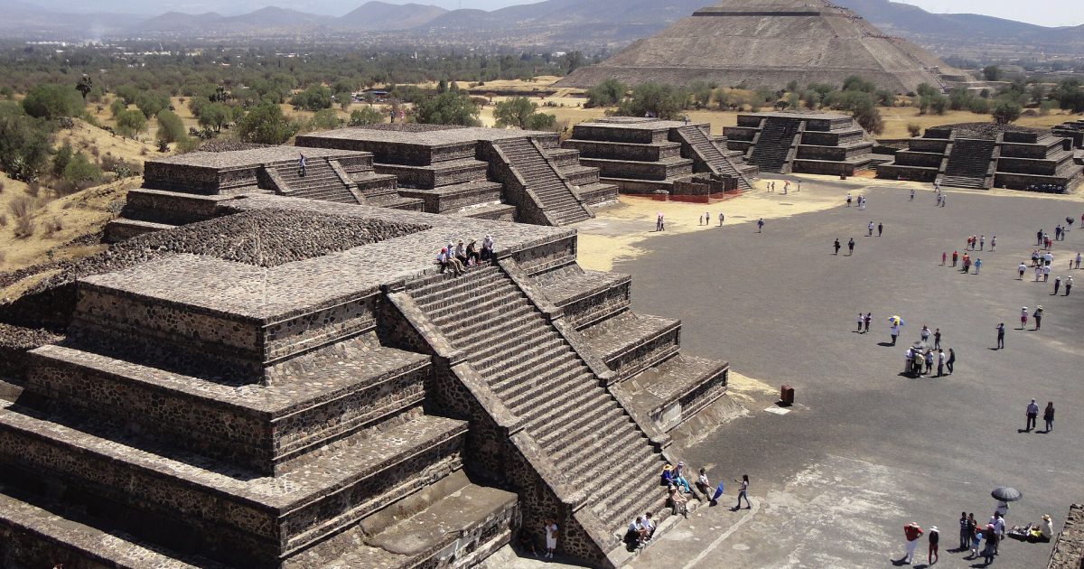 Aerial view of the ancient Teotihuacan pyramids, showcasing the Pyramid of the Moon and visitors exploring the site surrounded by mountains and greenery.