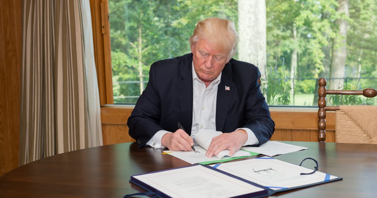 Donald Trump signing documents at a table with a view of greenery in the background.