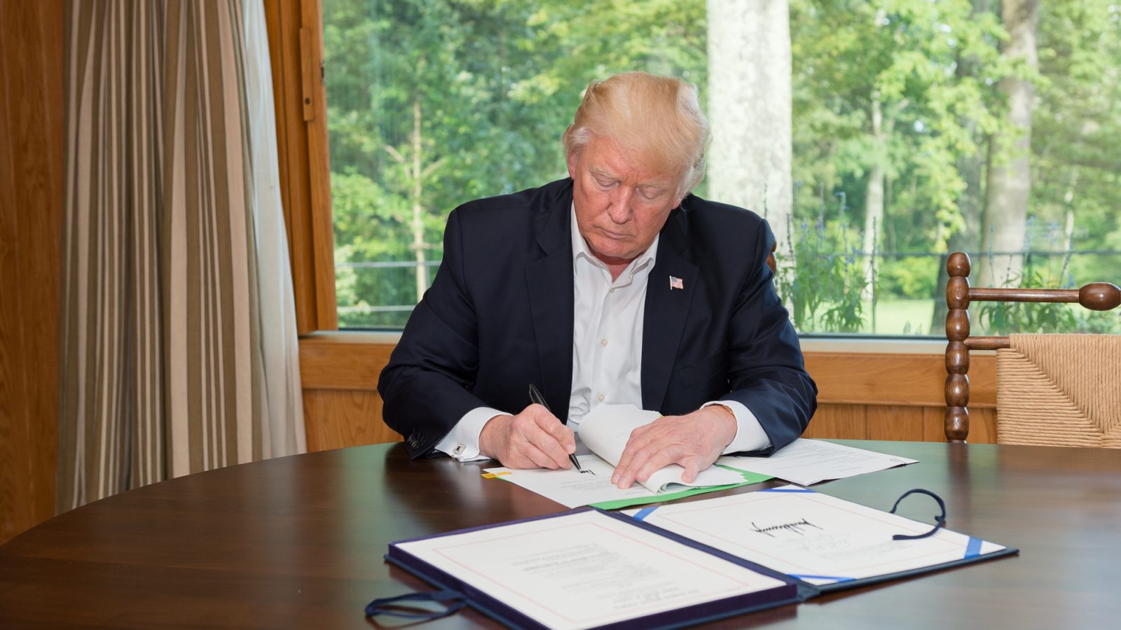 Donald Trump signing documents at a table with a view of greenery in the background.