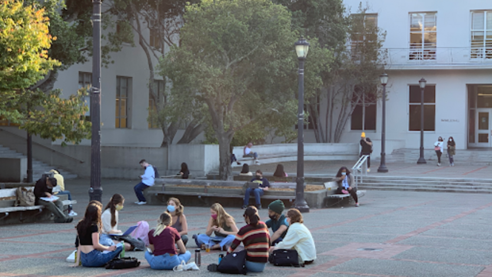 Students gather in a campus courtyard, sitting on the ground and engaging in discussion, with some individuals studying in the background.
