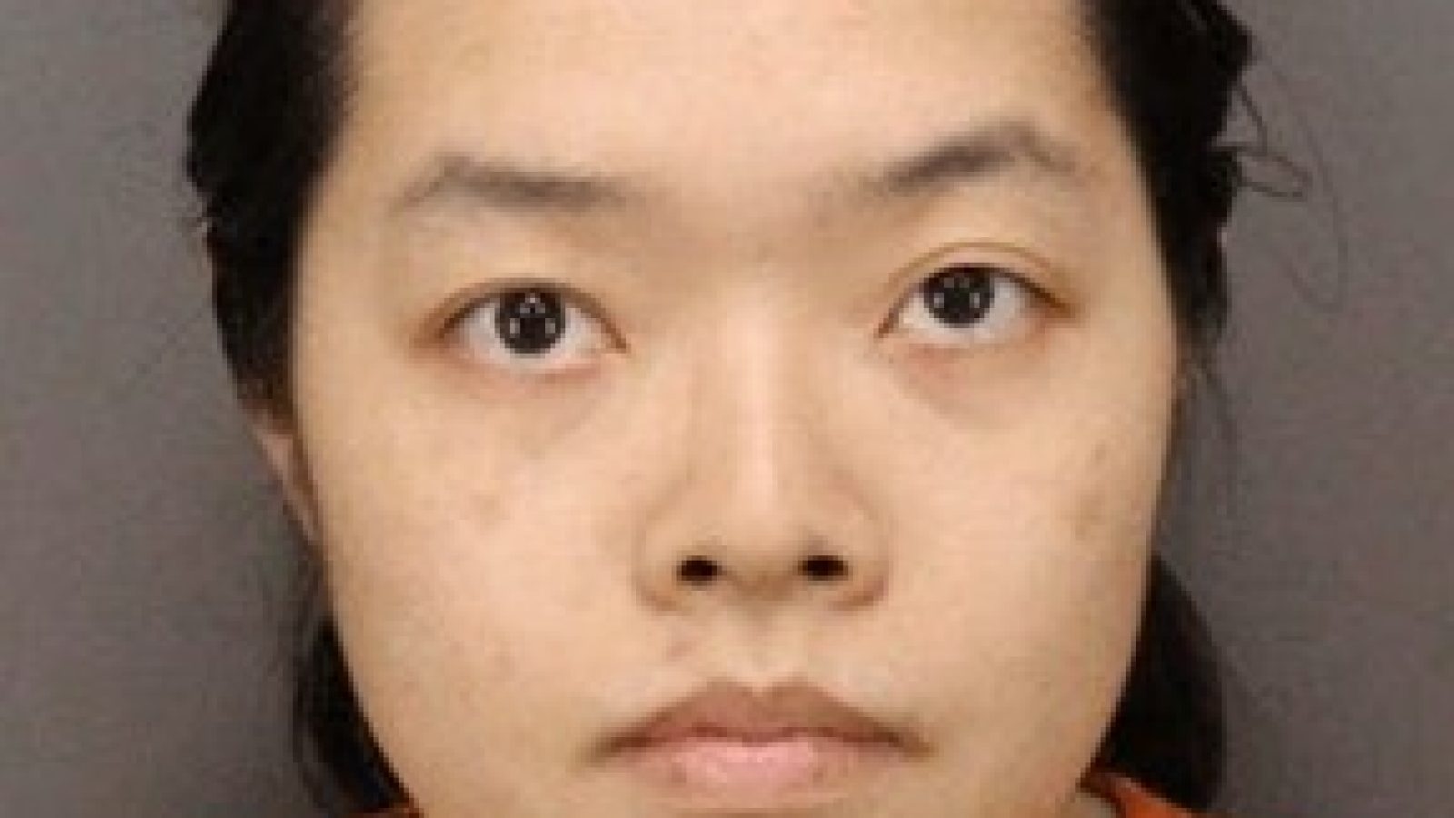 Mugshot of an individual with dark hair wearing an orange shirt, displaying a neutral expression against a gray background.