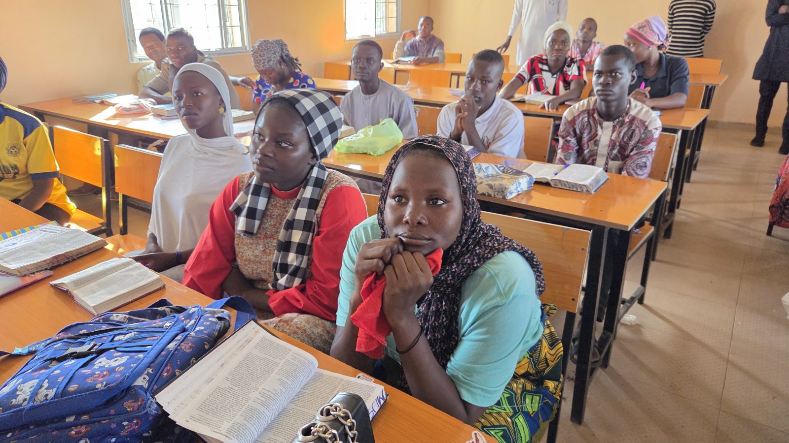 Students attentively listen in a classroom, surrounded by books and notebooks, showcasing a focused learning environment.