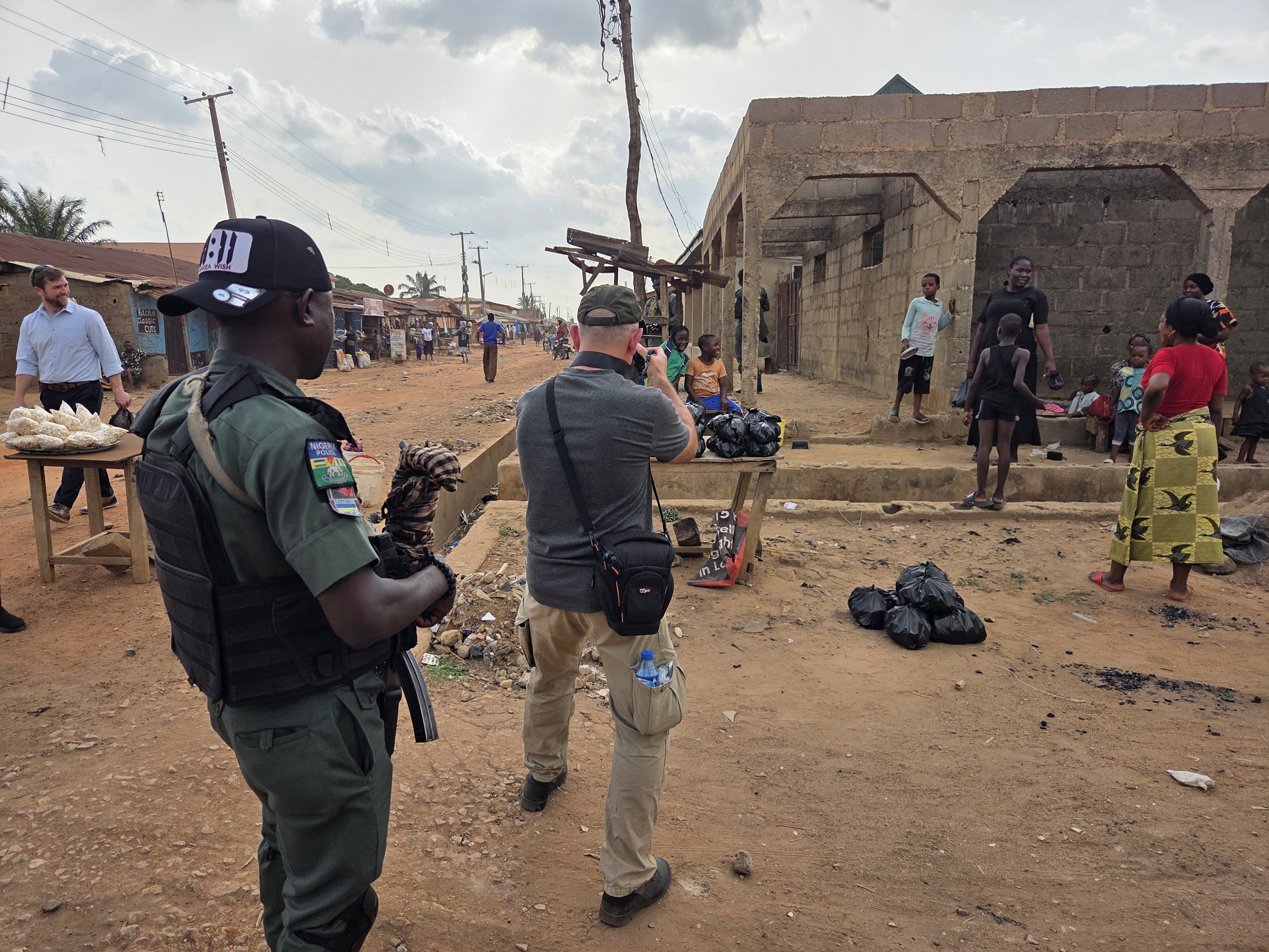 A security officer stands in a bustling market street while visitors explore local shops and interact with residents in a vibrant community setting.