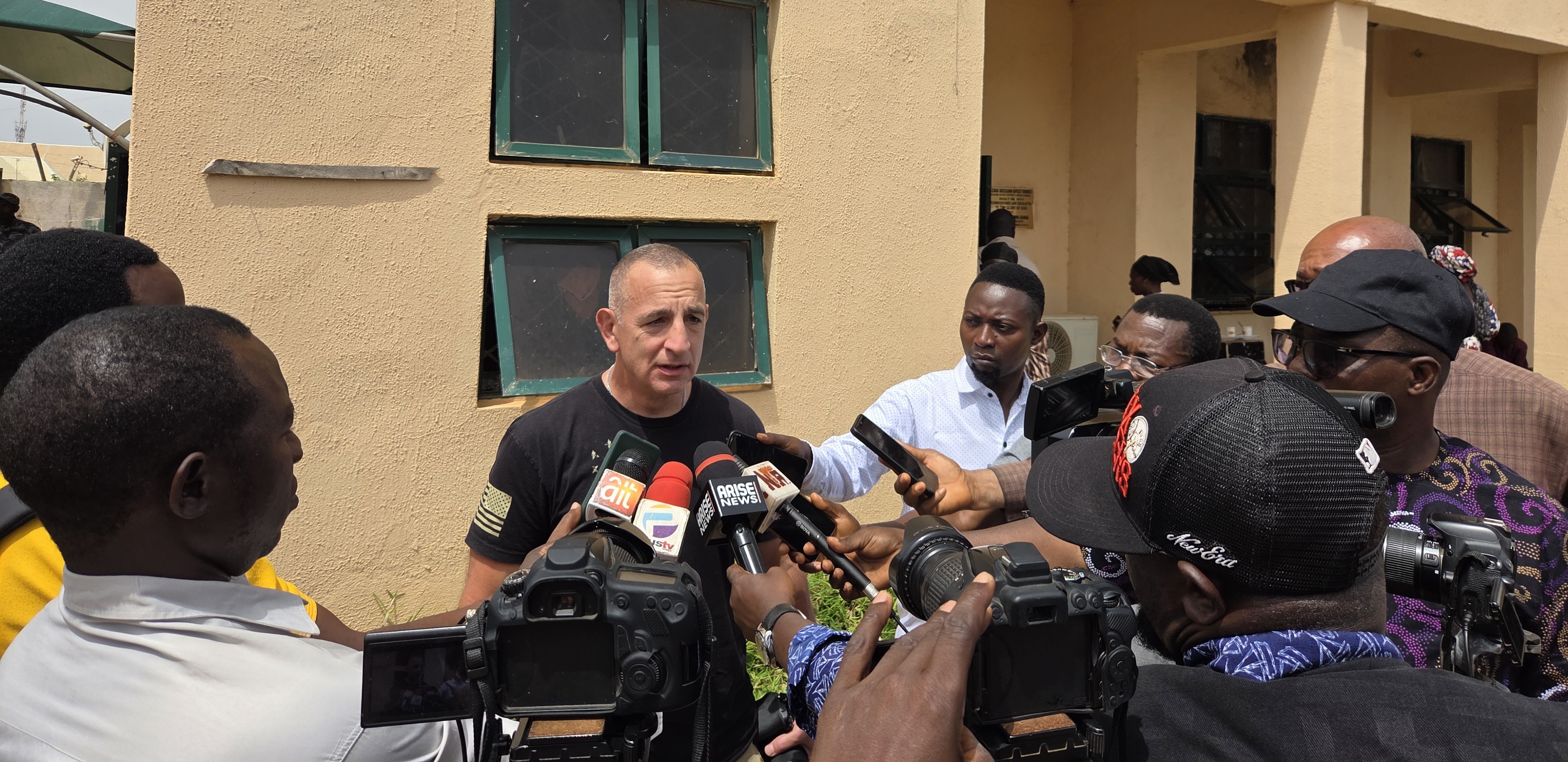 A speaker addresses a crowd of journalists and cameras outside a building, highlighting media engagement and press coverage.
