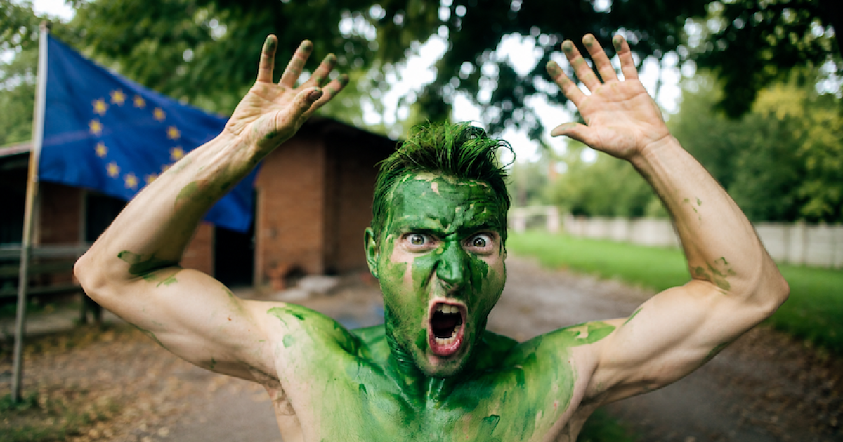 A man with green body paint and an intense expression poses outdoors, with an EU flag in the background.