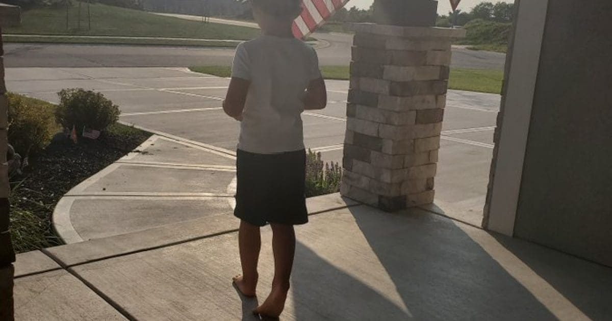A barefoot child stands on a porch, facing a driveway with American flags in the background during a sunny day.