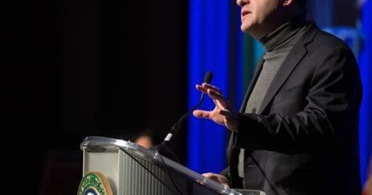 Speaker delivering a presentation at a podium with a microphone, emphasizing key points during an event with a blue backdrop.