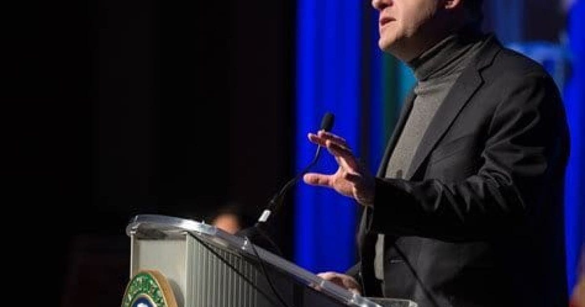 Speaker delivering a presentation at a podium with a microphone, emphasizing key points during an event with a blue backdrop.