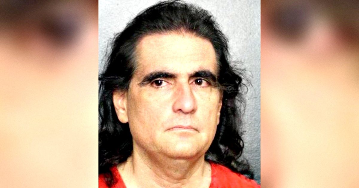 Mugshot of a man with long dark hair wearing a red shirt, showing a serious expression against a gray background.