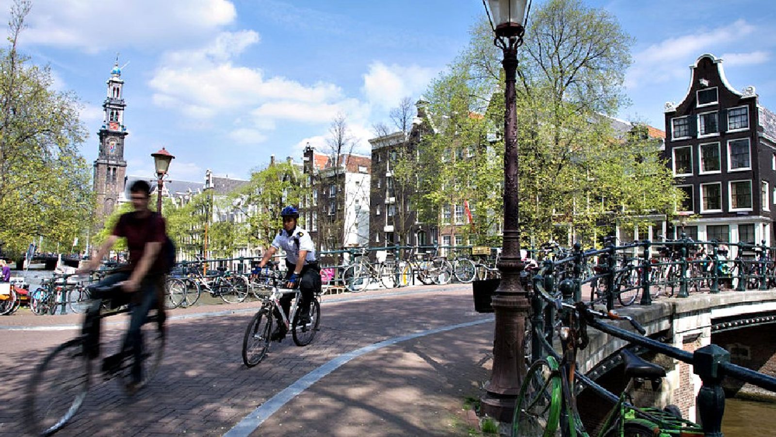 Bicyclists ride along a canal in Amsterdam with historic buildings and trees in the background under a clear blue sky.