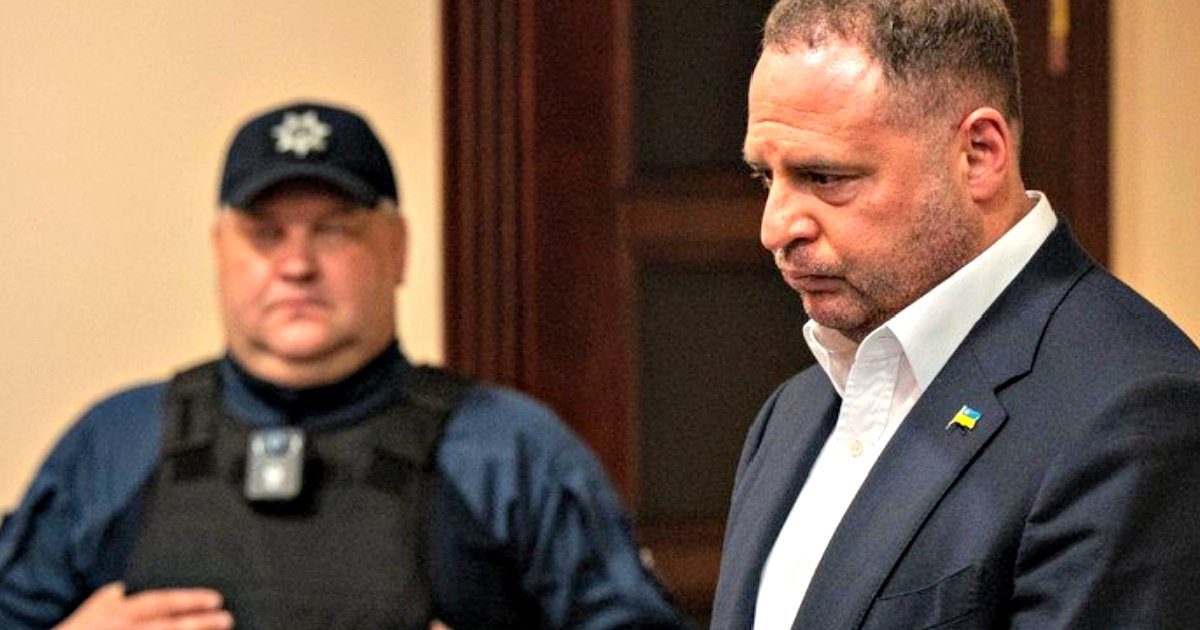 A serious man in a suit stands in a courtroom setting, accompanied by a police officer in uniform, highlighting a tense legal situation.