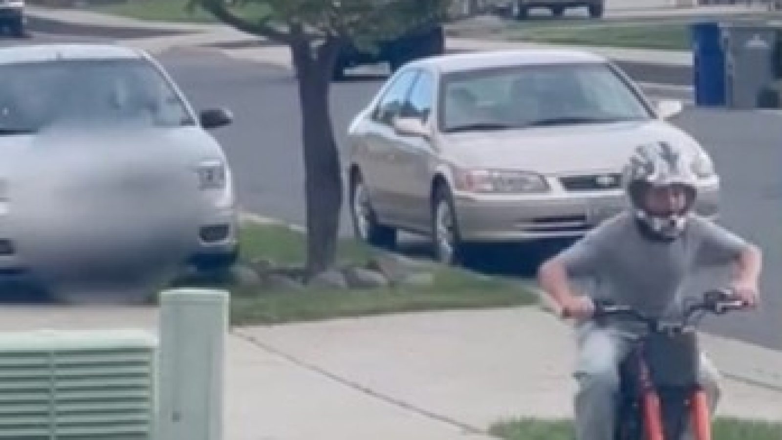 Child riding an electric bike on a suburban street, wearing a helmet, with parked cars and a tree in the background.