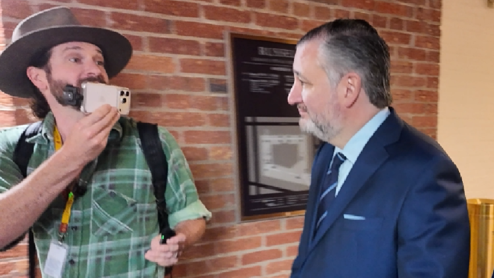 A man in a green plaid shirt and hat holds a phone while conversing with a suited politician in a brick hallway.