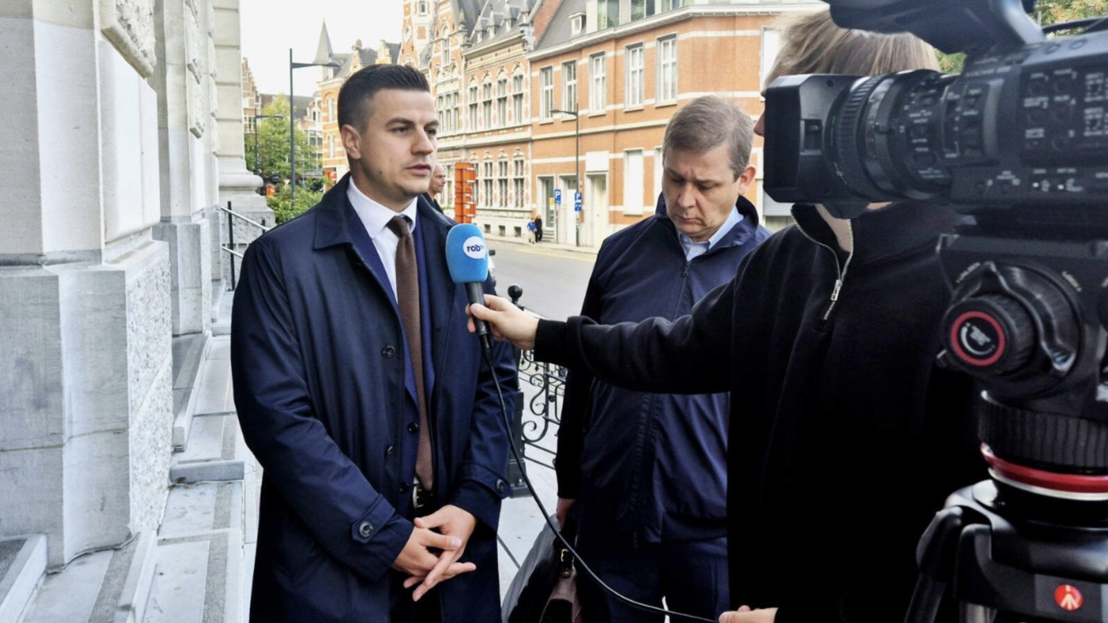 Two men, one being interviewed and the other observing, stand outside a building while a reporter conducts a video interview with a camera present.