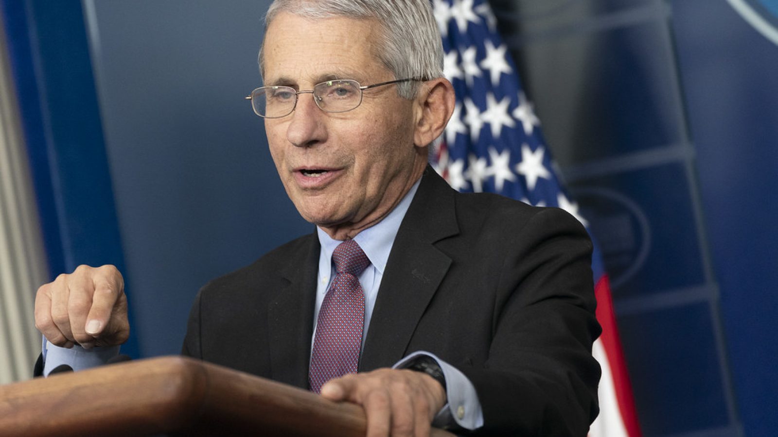 Dr. Anthony Fauci speaks at a podium in a press briefing, with the American flag in the background.