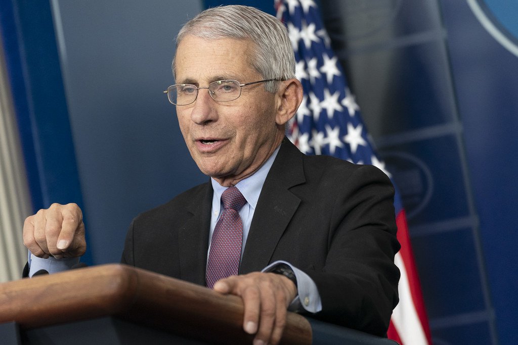 Dr. Anthony Fauci speaks at a podium in a press briefing, with the American flag in the background.