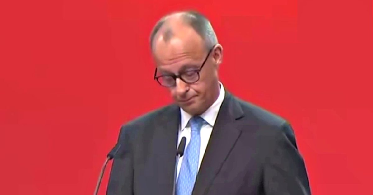 Man in a suit and glasses speaking at a podium against a red background, displaying a serious expression during a public address.