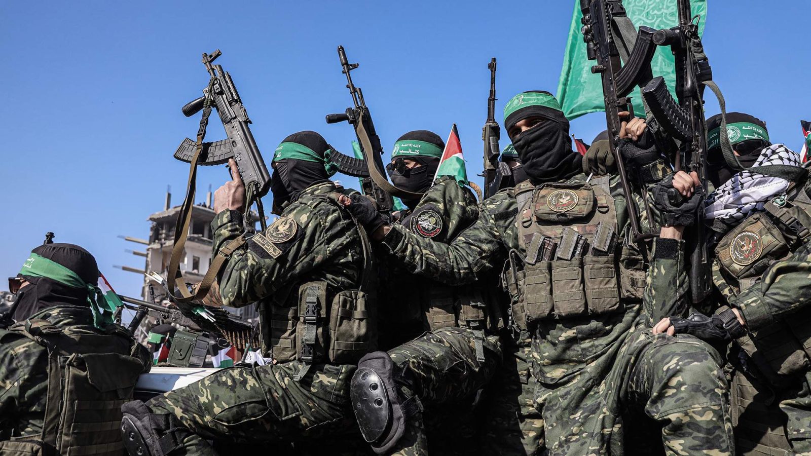 Militant group members in camouflage uniforms display weapons during a demonstration, with flags in the background under a clear blue sky.