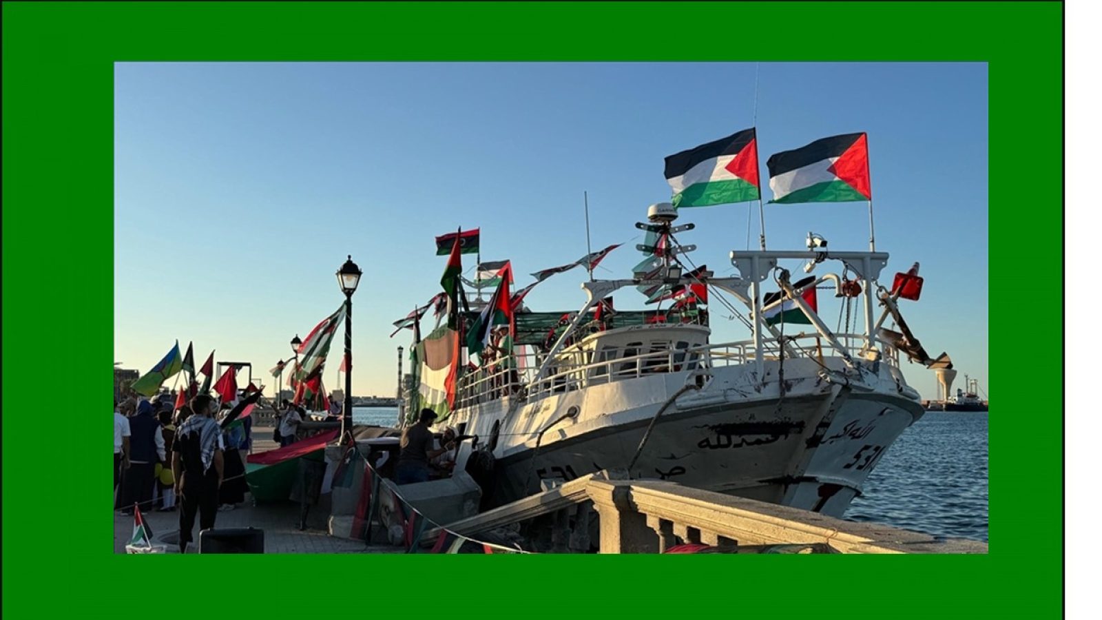 A decorated boat with Palestinian flags is docked at a waterfront, surrounded by people and colorful banners during a cultural or political event.