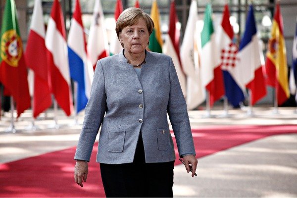 Angela Merkel walks on a red carpet surrounded by international flags, symbolizing her leadership and diplomatic presence in global politics.
