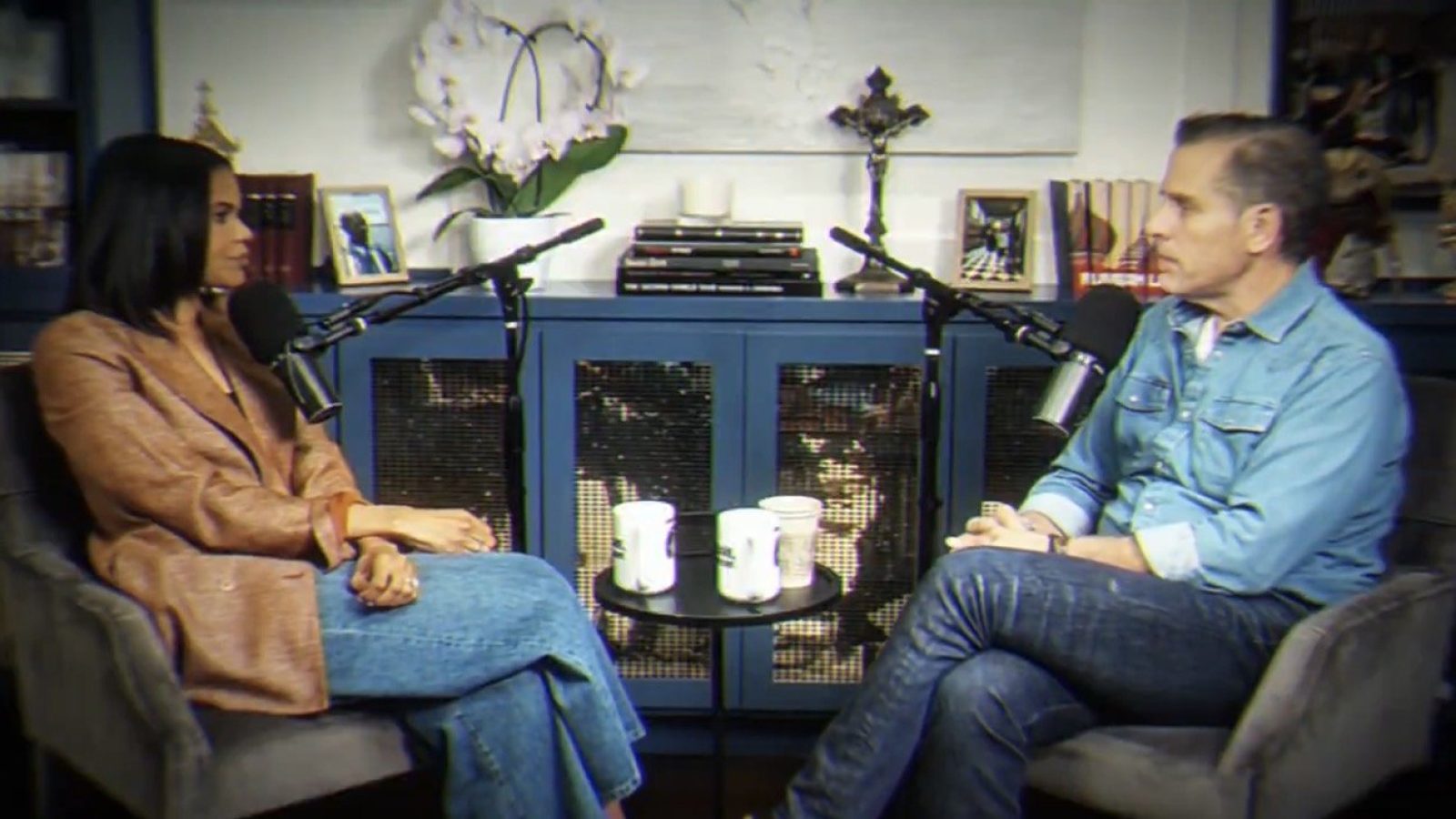 A woman and a man engage in a conversation during a podcast recording, seated in a cozy studio with microphones and coffee cups on a small table.