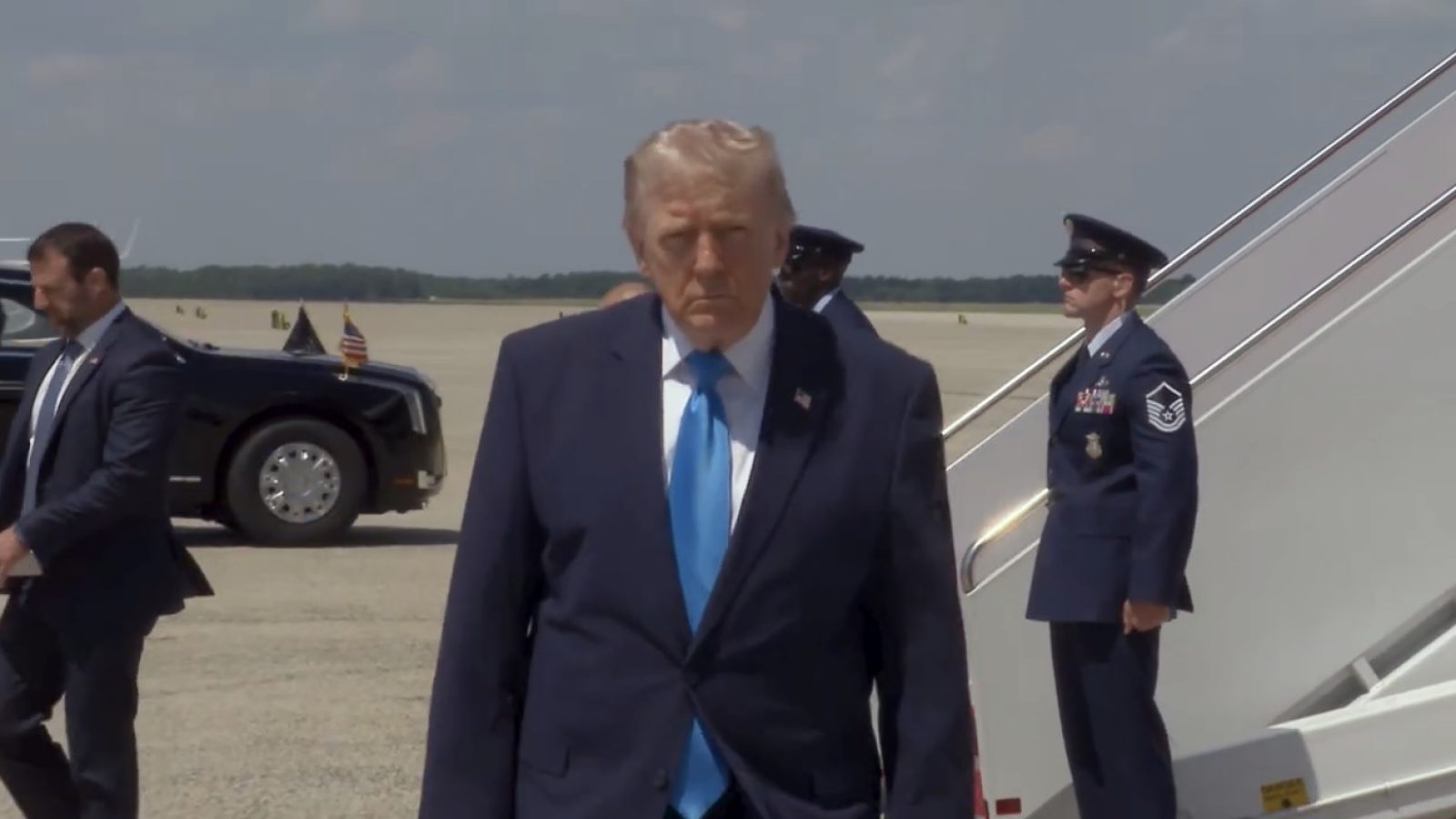 Former President Donald Trump walks towards Air Force One on a tarmac, flanked by security personnel and military members.