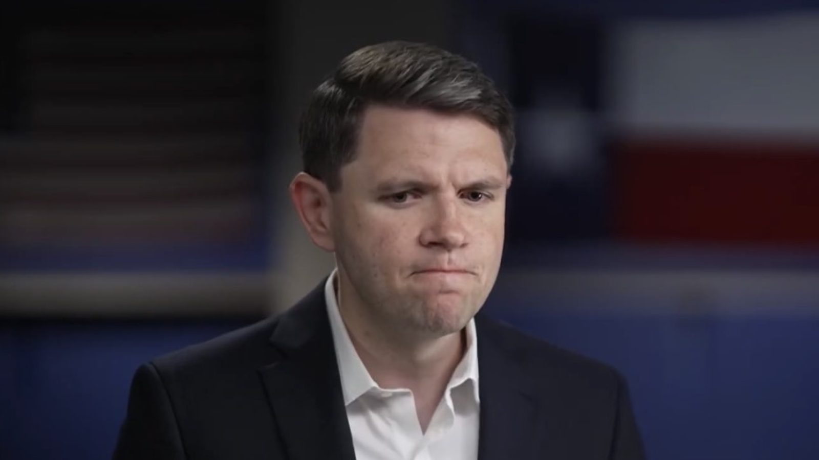 Man in a suit with a concerned expression, sitting in front of a blurred Texas flag background.