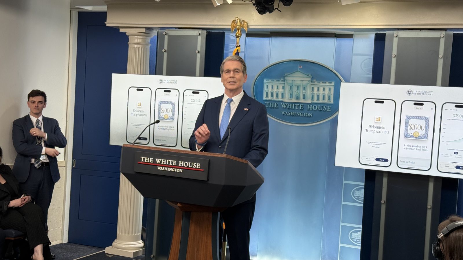Official White House press conference featuring a speaker discussing Trump Accounts, with informational graphics displayed on screens in the background.