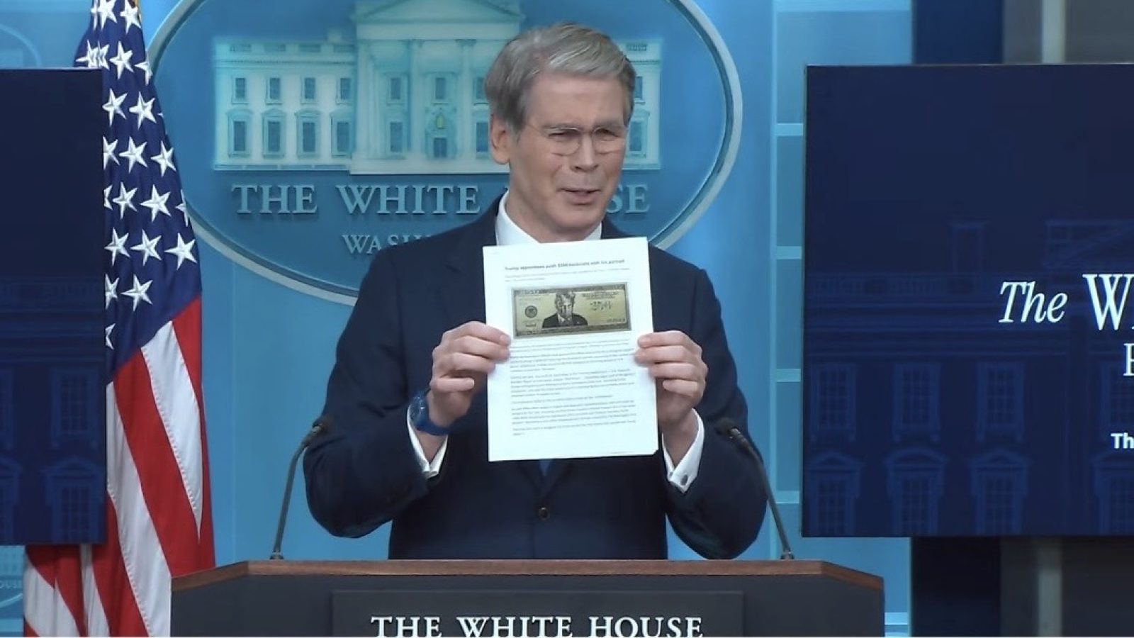 Official at the White House briefing holds a document and a bill, discussing important financial information with the American flag in the background.