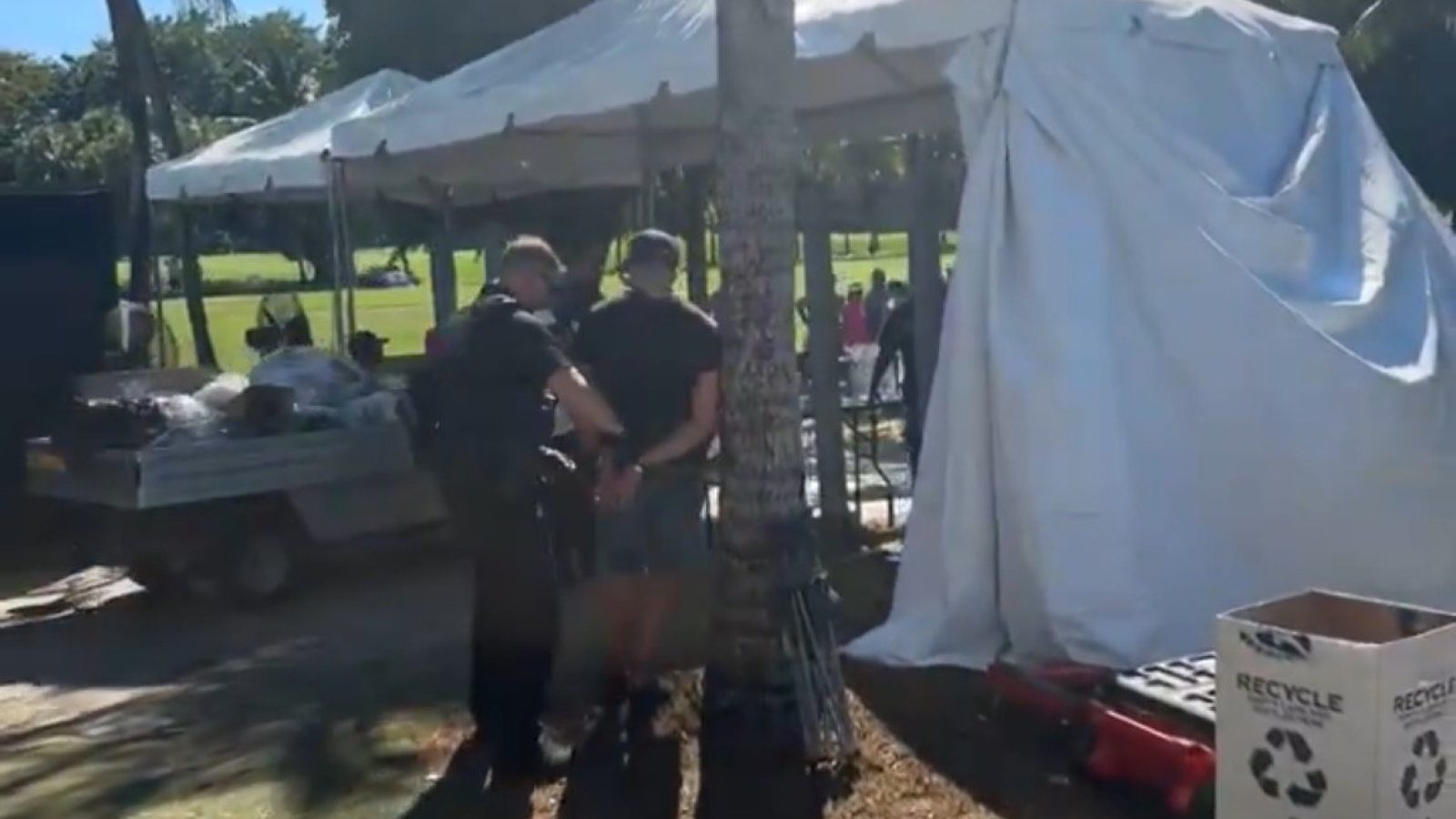 Police officer apprehending a suspect near a tent at an outdoor event, with a crowd in the background and equipment nearby.