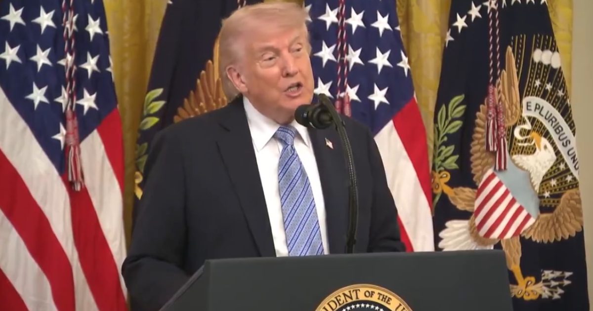 Donald Trump speaking at a podium in front of U.S. flags and presidential seals during a press conference.