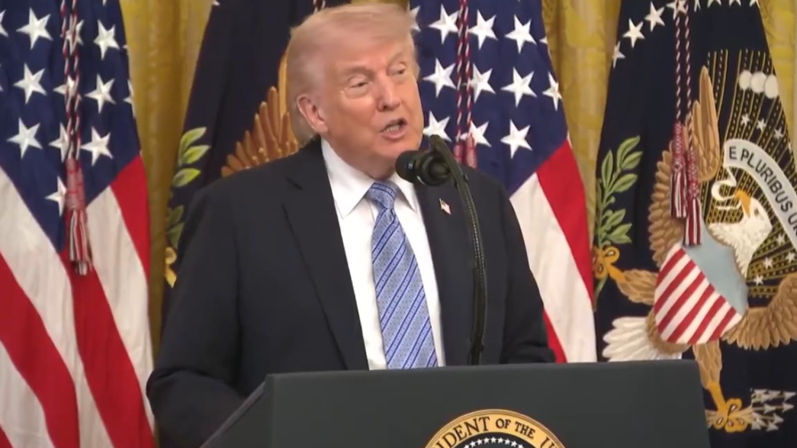 Donald Trump speaking at a podium in front of U.S. flags and presidential seals during a press conference.